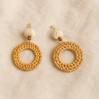 Pair of round hand-woven rattan earrings with natural fiber beads on beige fabric background.