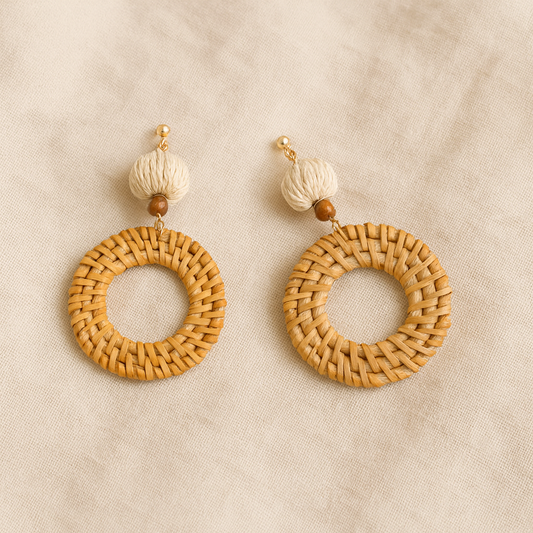 Pair of round hand-woven rattan earrings with natural fiber beads on beige fabric background.