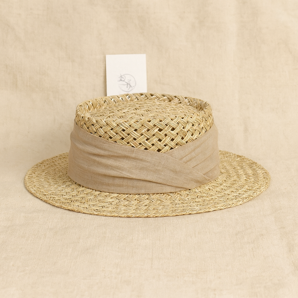 Natural woven straw hat with a beige linen ribbon, placed on linen fabric background showcasing craftsmanship details.