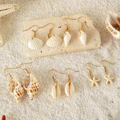 Collection of handmade shell earrings with gold hooks arranged on sand, featuring starfish, cowrie, and spiral designs.