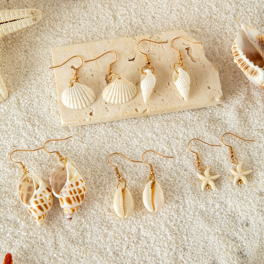 Collection of handmade shell earrings with gold hooks arranged on sand, featuring starfish, cowrie, and spiral designs.