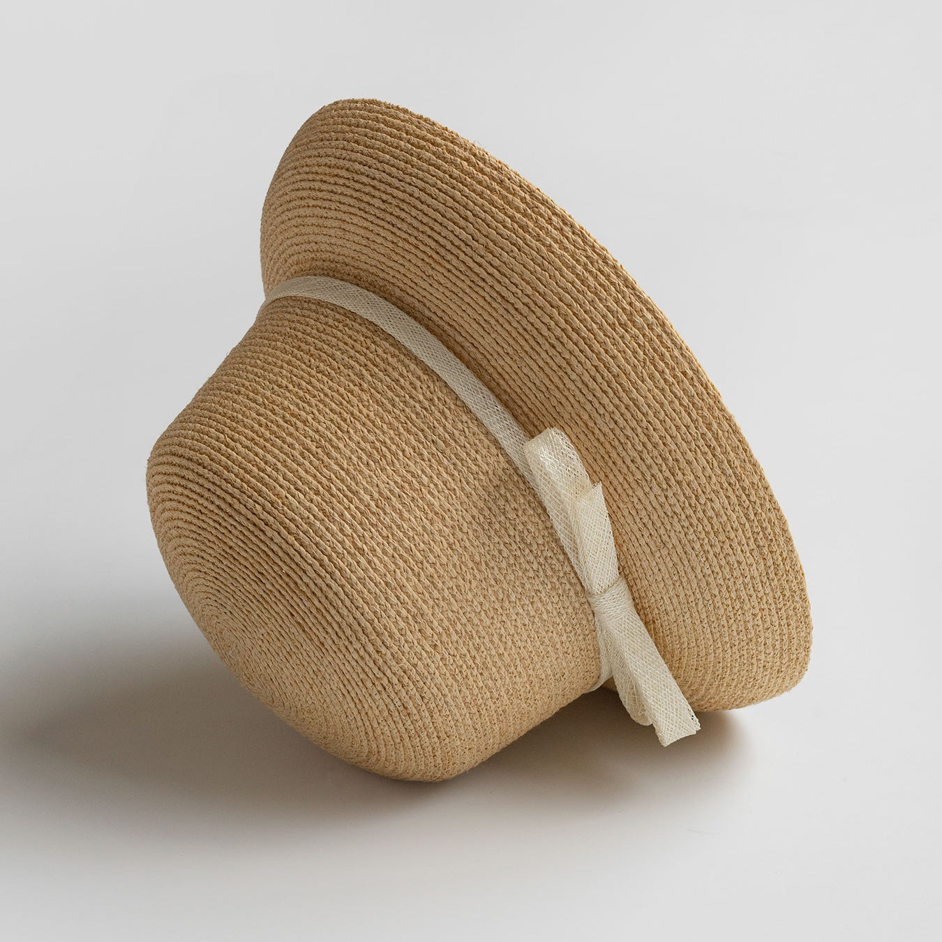 Beige woven straw hat with cream bow, viewed from side angle on light background to show shape and texture.