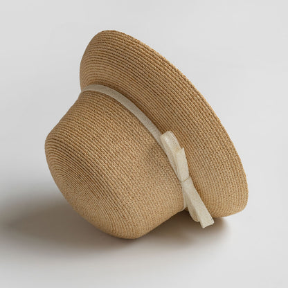 Beige woven straw hat with cream bow, viewed from side angle on light background to show shape and texture.