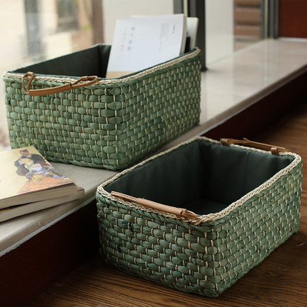 Two rectangular green woven straw baskets with leather handles placed by a window, ideal for home organization and decoration.