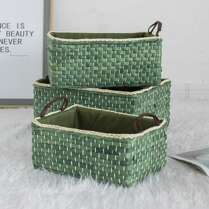 Set of three stackable green woven storage baskets with leather handles arranged vertically on a soft white rug.
