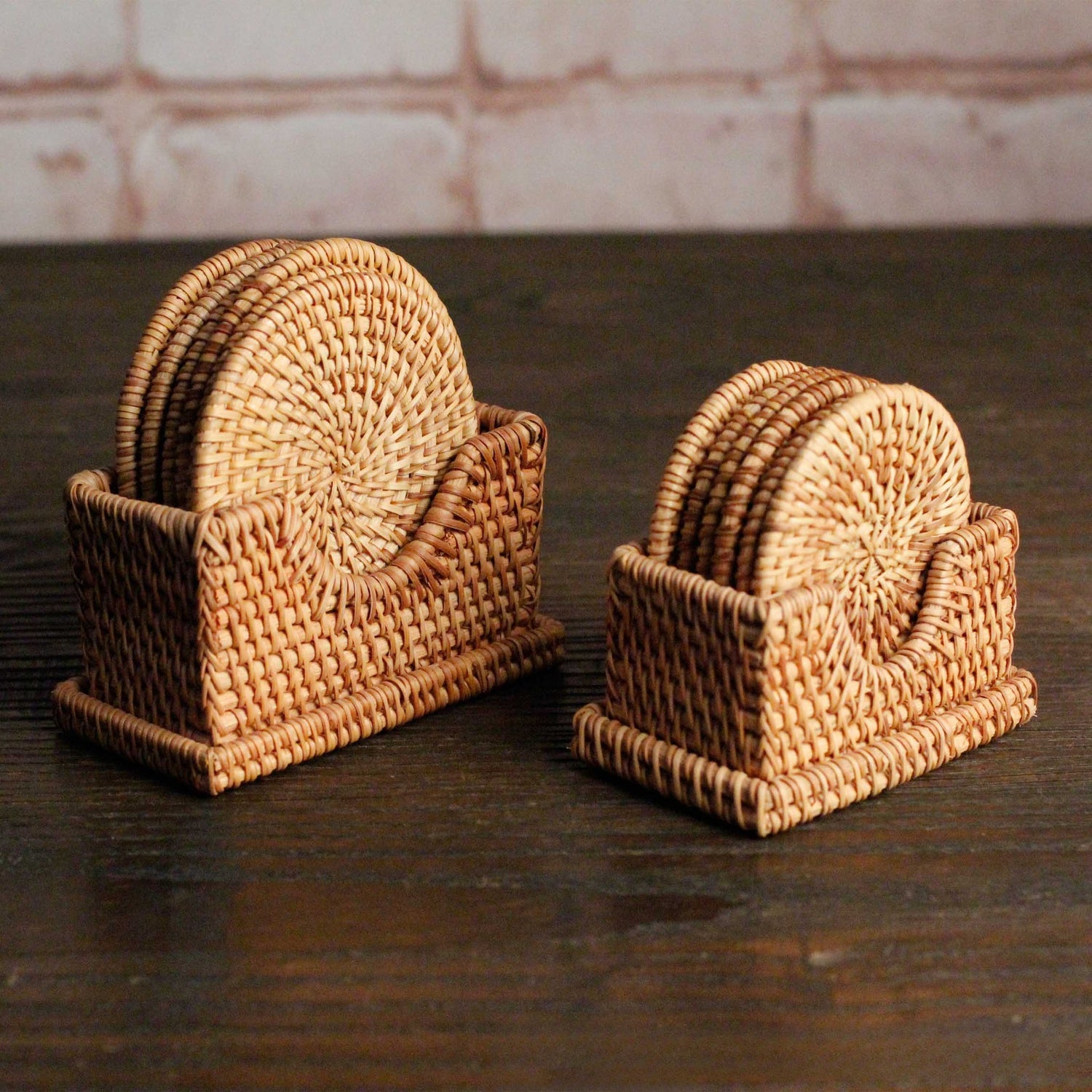 Two sizes of handwoven rattan coaster sets with holders placed side by side on dark wooden surface.