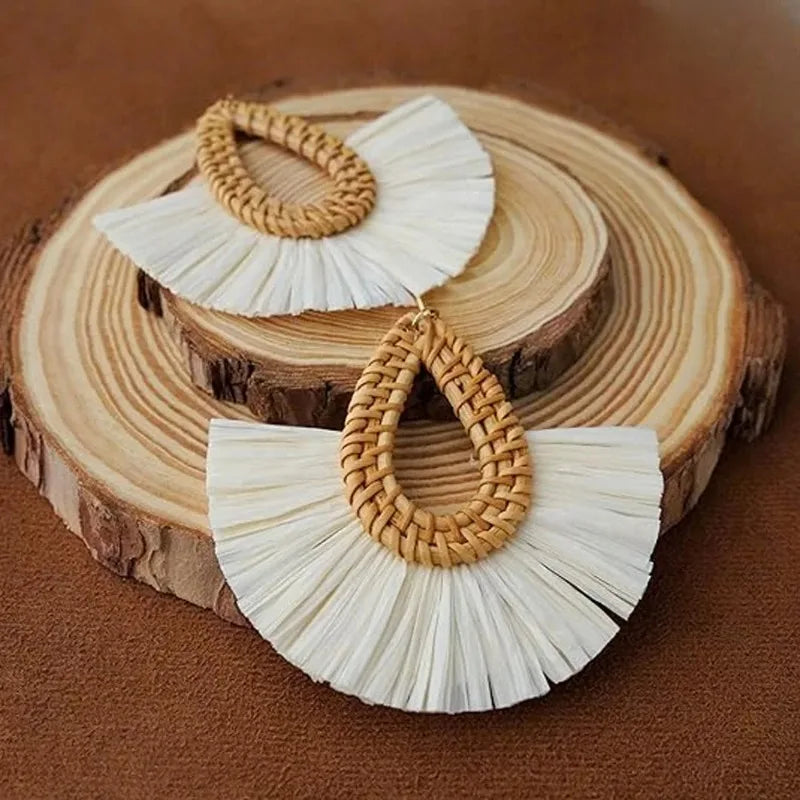 Handmade rattan teardrop earrings with white raffia fringe displayed on wooden round board.