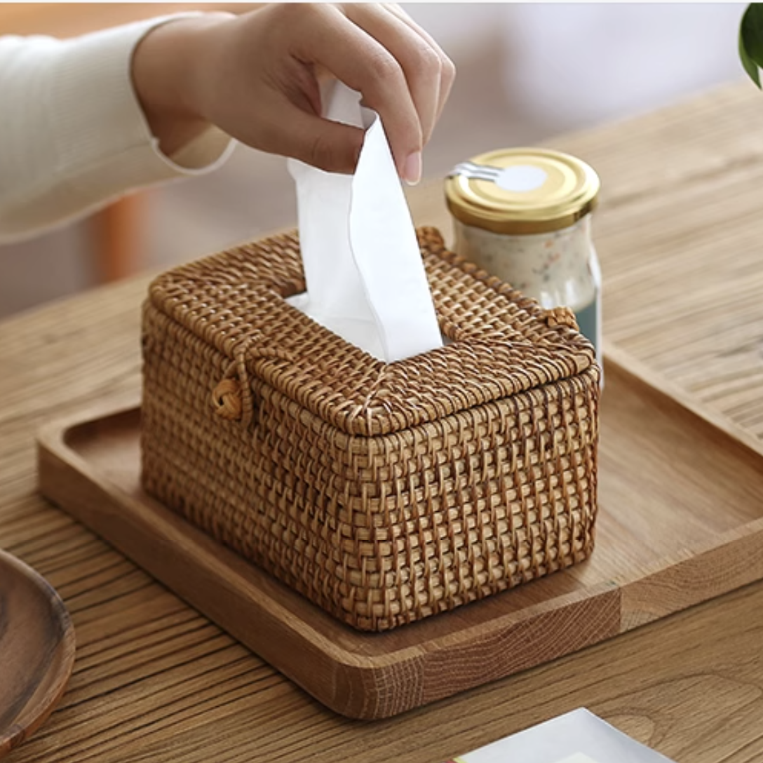 Hand pulling tissue from handmade rattan tissue box on wooden tray beside coffee and breakfast items.