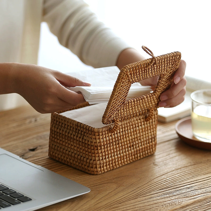 Person refilling handmade rattan tissue box by lifting lid and placing tissues inside on wooden table.