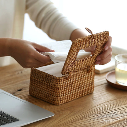 Person refilling handmade rattan tissue box by lifting lid and placing tissues inside on wooden table.