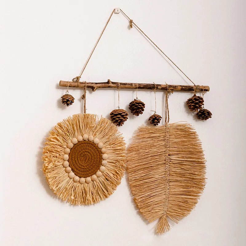 Raffia straw wall hanging with circular plate and leaf design, suspended from a wooden branch with pine cones.