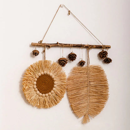 Raffia straw wall hanging with circular plate and leaf design, suspended from a wooden branch with pine cones.