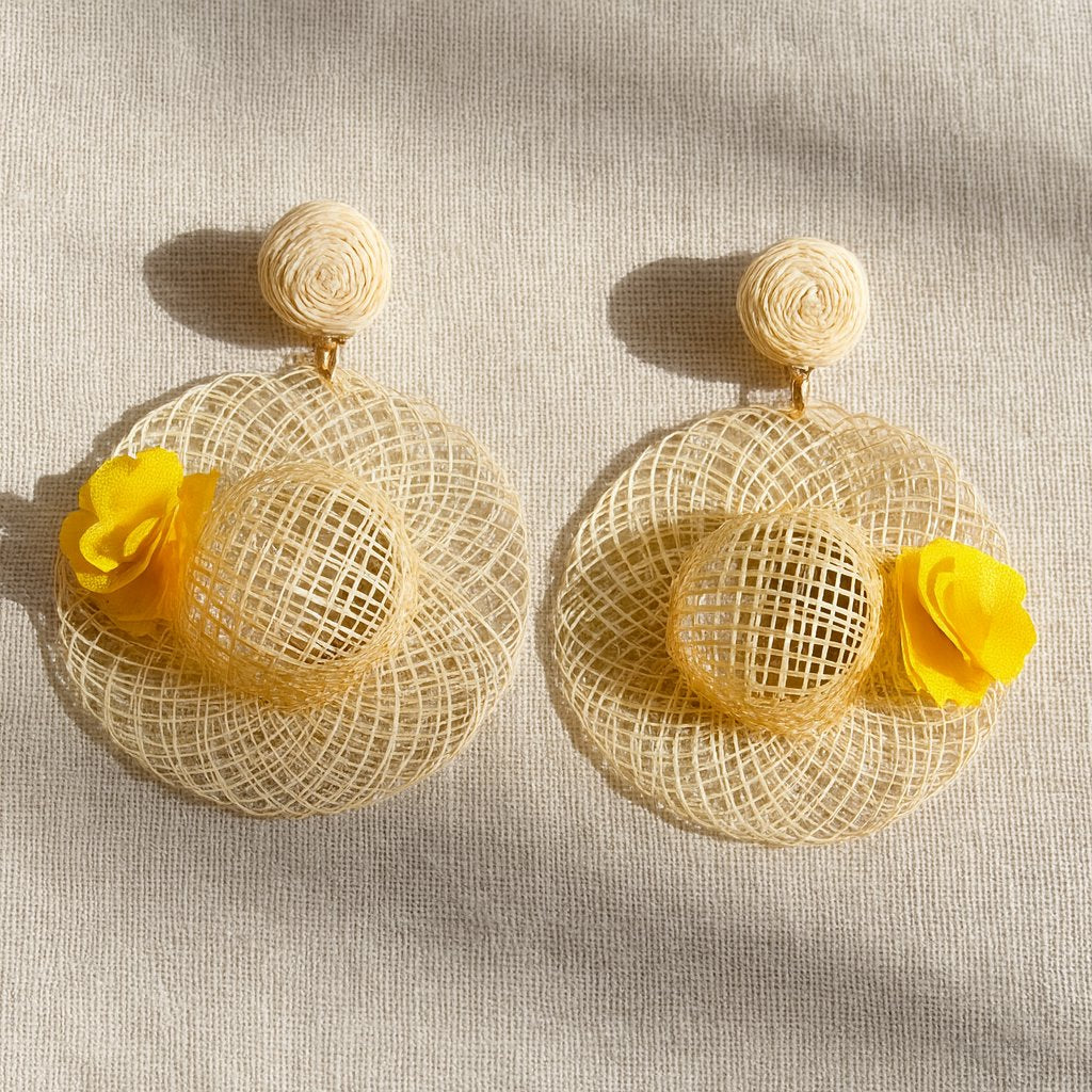 Handwoven straw hat earrings with yellow flower detail displayed on beige linen background.