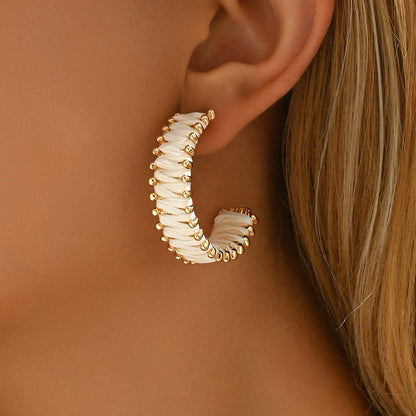 Close-up of single handwoven straw hoop earring with gold beads worn on ear.
