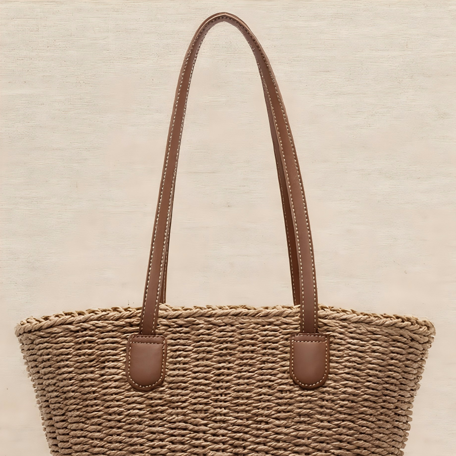 A brown handwoven straw tote with brown leather straps, displayed against a light neutral background.