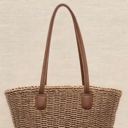 A brown handwoven straw tote with brown leather straps, displayed against a light neutral background.