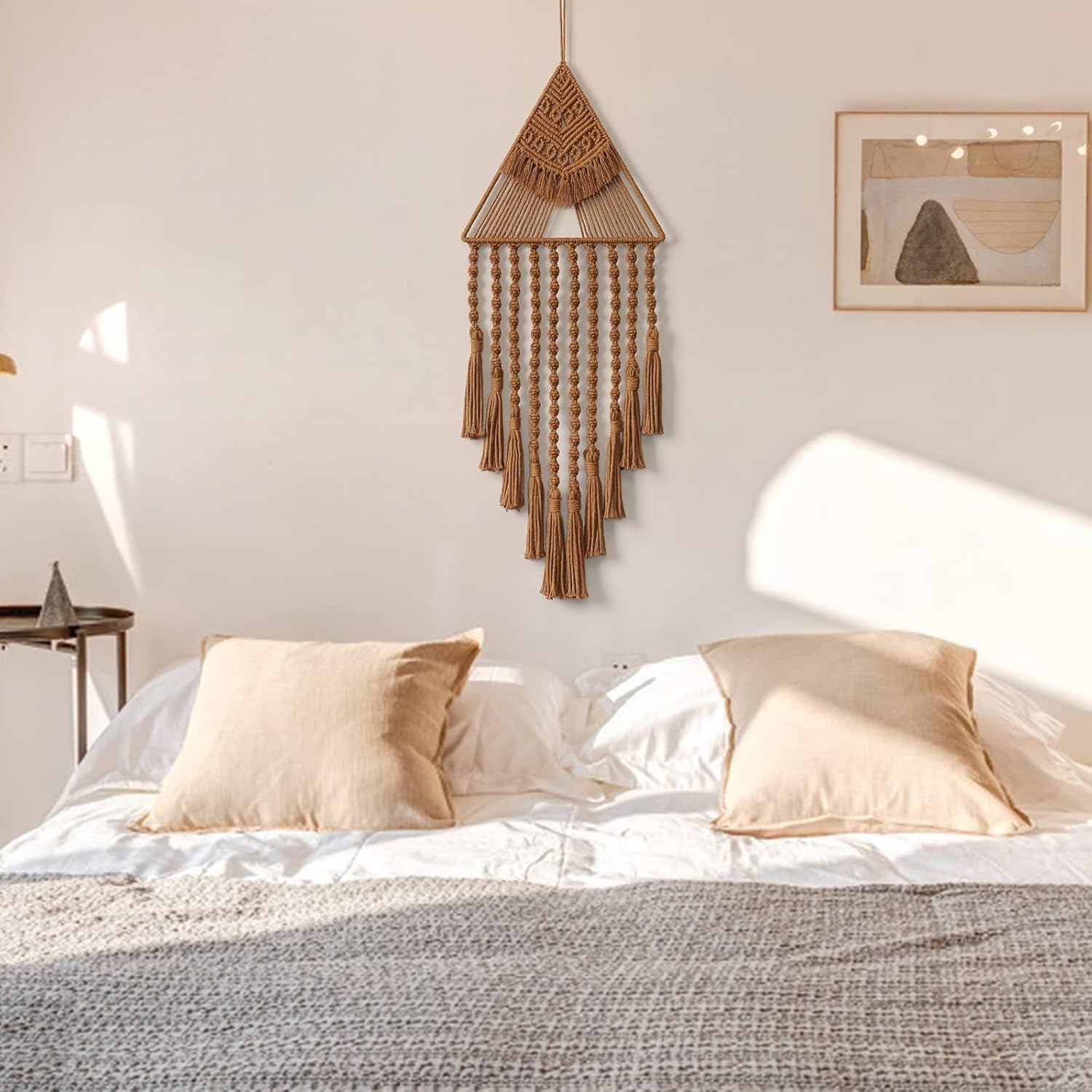 Bohemian macrame tapestry hanging above a minimalist bed with beige pillows and soft sunlight.