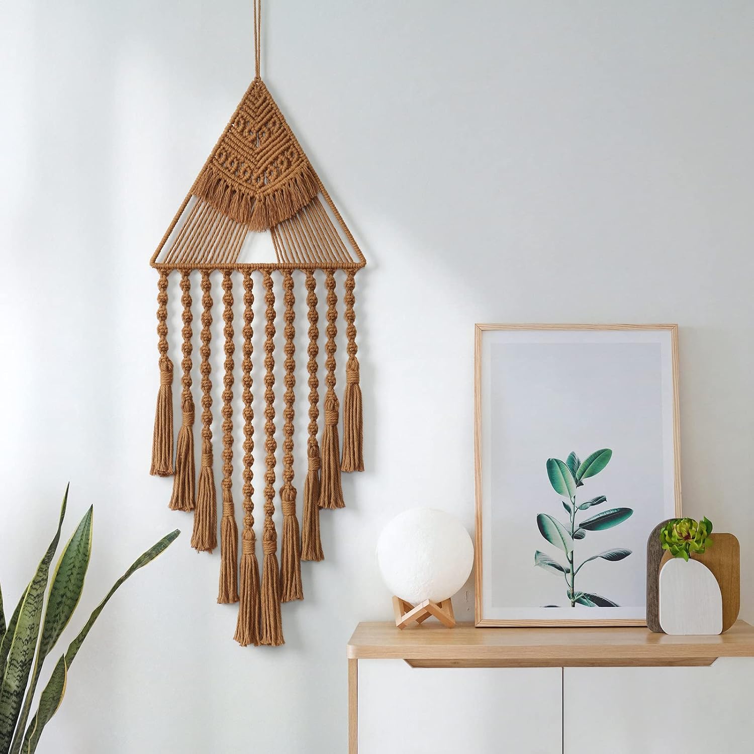 Handcrafted triangle macrame wall hanging displayed on white wall above modern sideboard and decor.