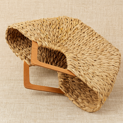 Angled view of the handwoven straw tote bag showing its curved structure and sturdy wooden handles on a beige textile background.