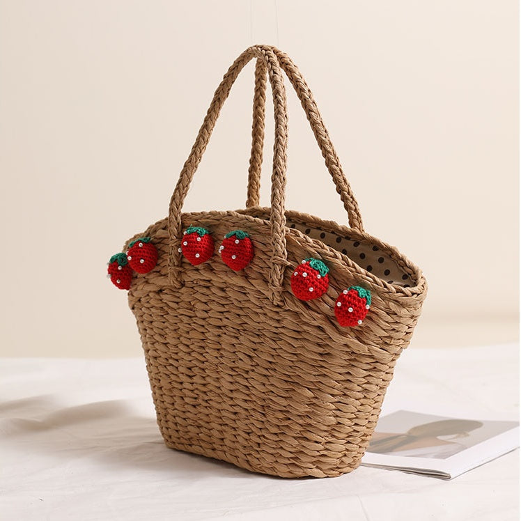 Dark straw tote bag with red crochet strawberries viewed from a slight angle, showing braided handles and dotted inner lining.