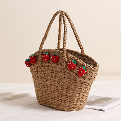 Dark straw tote bag with red crochet strawberries viewed from a slight angle, showing braided handles and dotted inner lining.