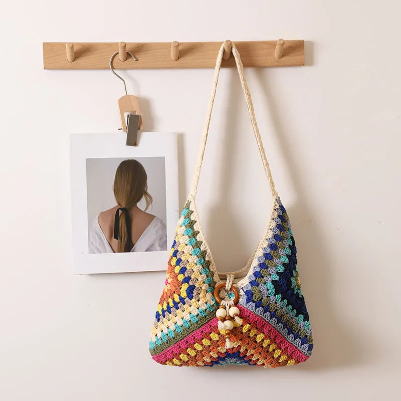 Multicolor crochet cotton shoulder bag with diagonal rainbow pattern, hanging on wooden hook beside a minimalist photo print.