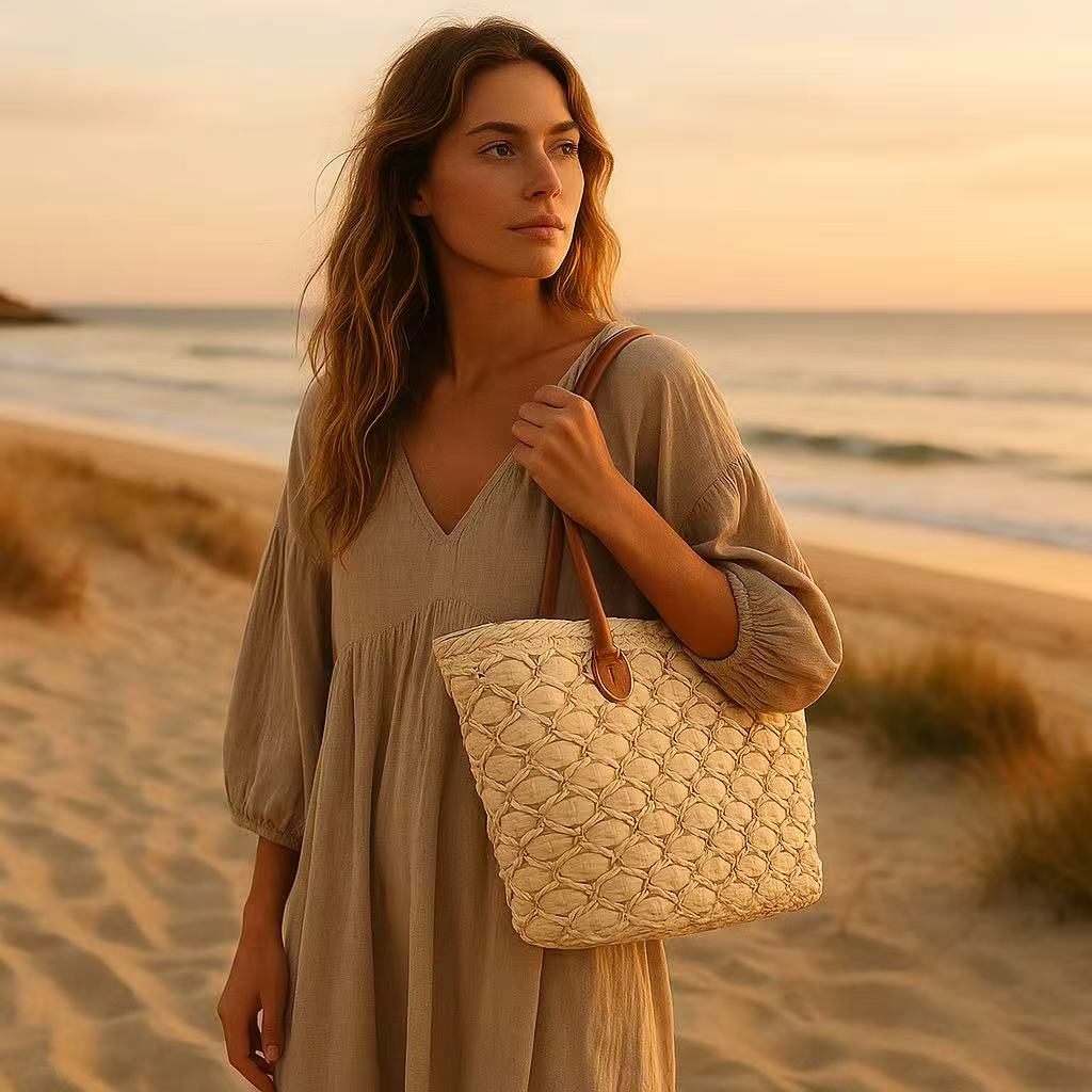 Woman in a beige dress holding a woven straw tote bag with brown handles while walking on the beach at sunset.
