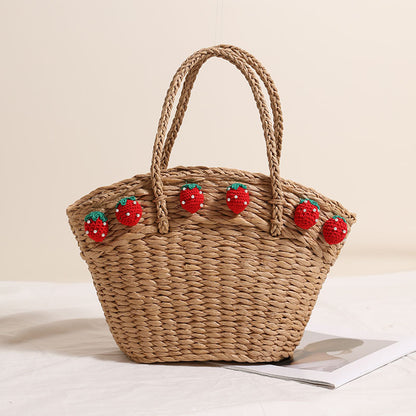 Dark brown straw beach tote bag with red crochet strawberry decorations along the top edge, shown upright on a white surface.