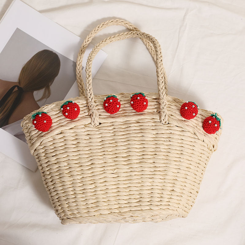 Light straw tote bag with strawberry decorations placed flat beside a minimalist photo print, showing woven texture and curved silhouette.