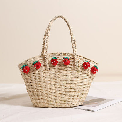 Light-colored straw beach tote bag decorated with crochet strawberries, placed on a soft white background beside an open magazine.