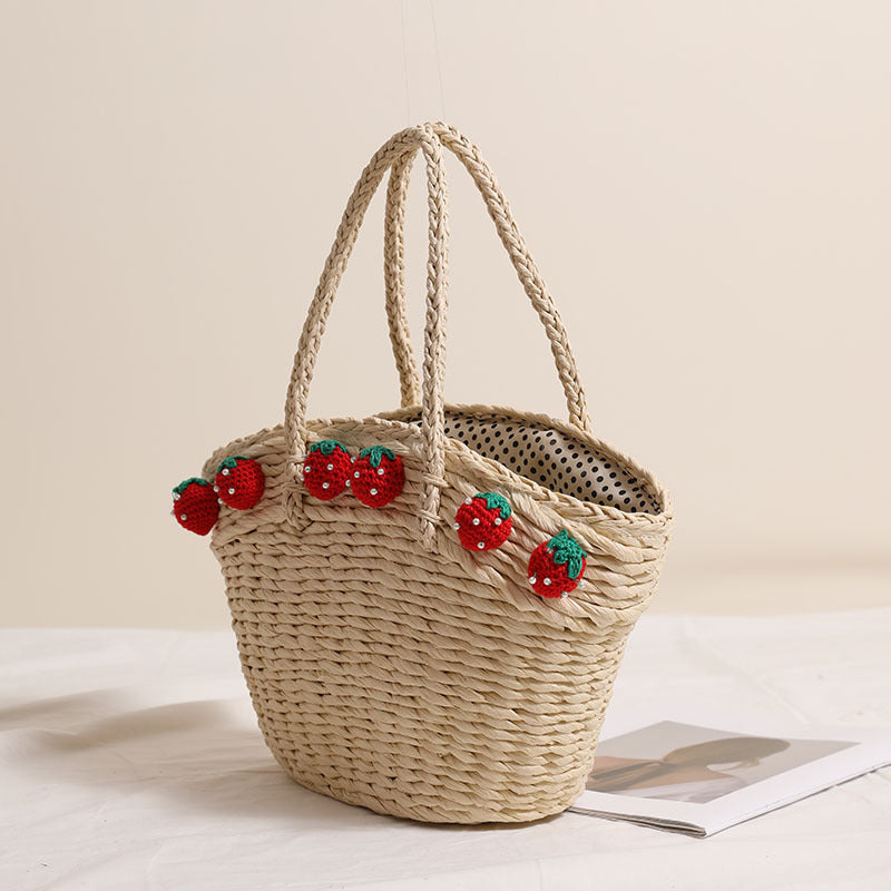 Light straw tote bag with crochet strawberry charms, viewed from an angle to show its spacious shape and inner lining.