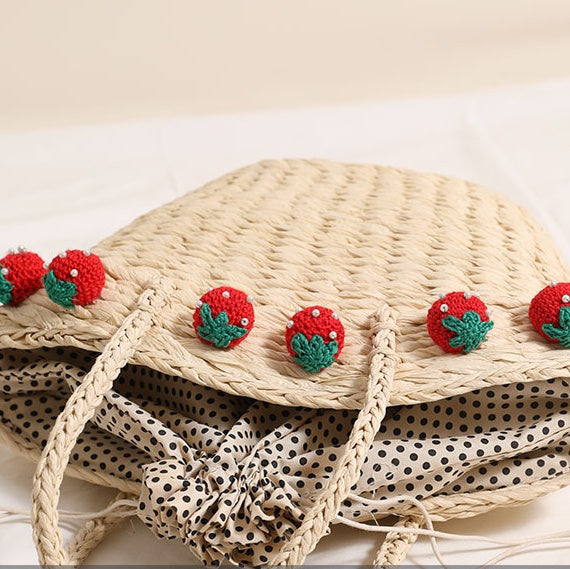 Close-up of the drawstring interior and polka-dot lining of the light straw tote bag with red crochet strawberry charms.