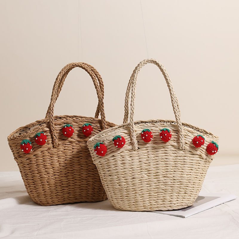 Pair of handwoven straw tote bags, one light and one dark, each adorned with red crochet strawberries, displayed together on a white cloth.