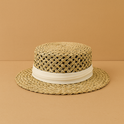 Handwoven flax straw hat with white ribbon accent displayed on a warm beige background, showing its natural woven texture.