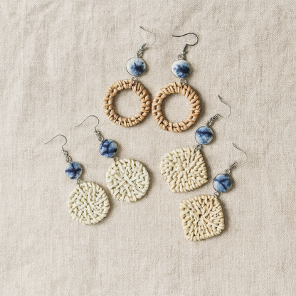 Set of handmade vine and straw earrings in round and square shapes with blue ceramic beads displayed on linen fabric.