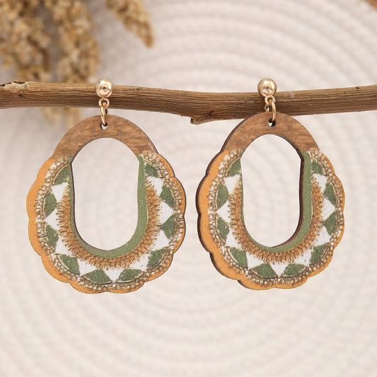 Natural wooden earrings with green and white embroidered geometric pattern hanging on a branch.