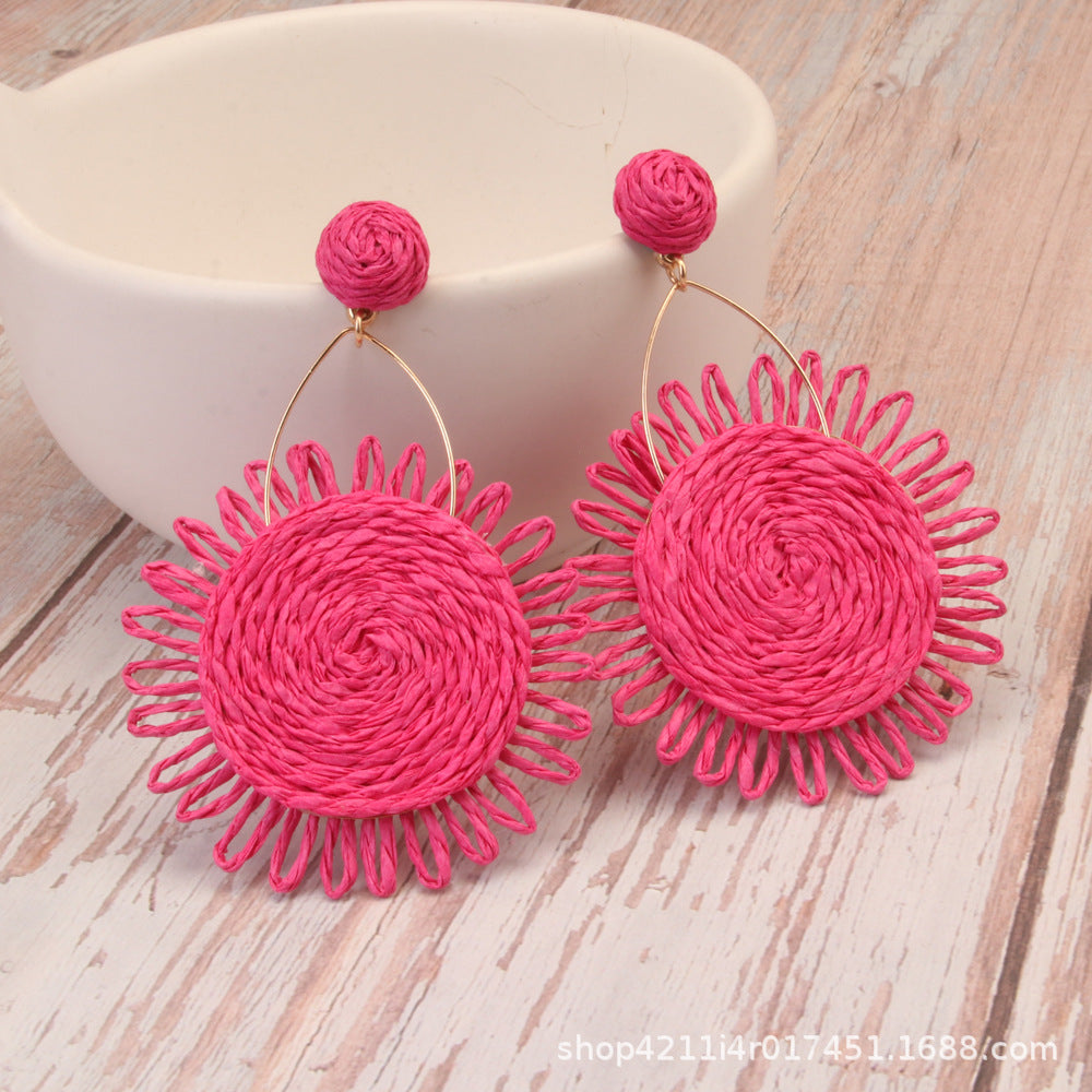 Pink woven Lafite grass sunflower earrings with circular raffia design on wooden background.