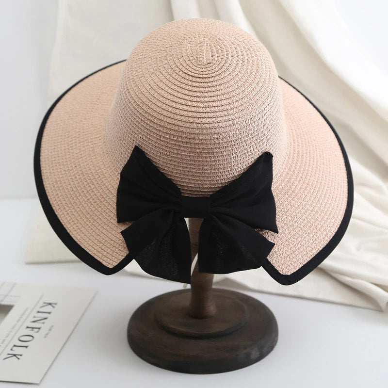 Soft pink-toned straw hat with black ribbon bow, shown on wooden base against white fabric background.