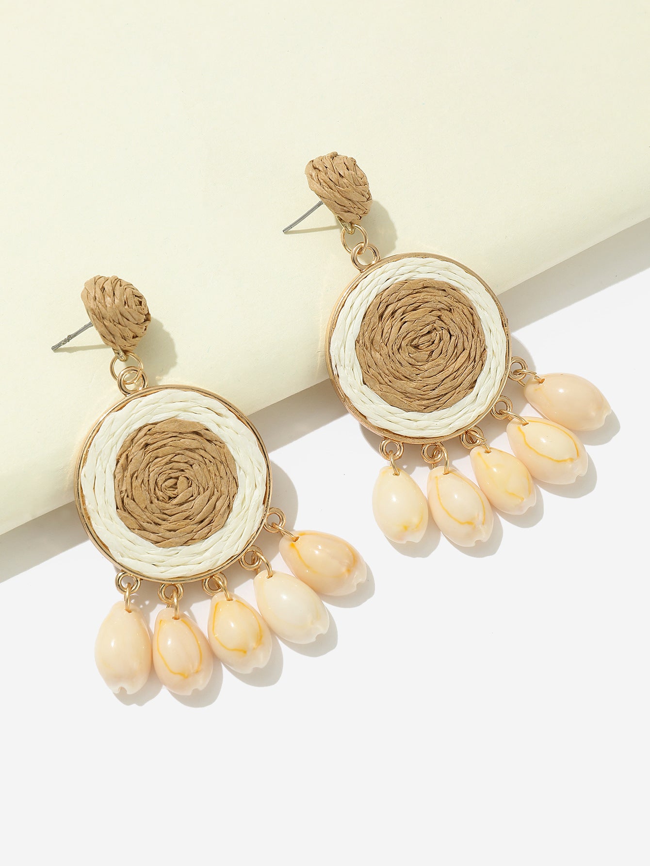 Beige raffia-woven shell earrings with layered shells photographed on bright white background.