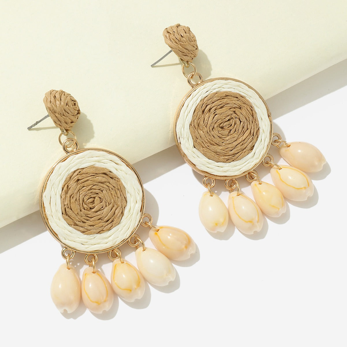 Beige raffia-woven shell earrings with gold frame displayed on white background.