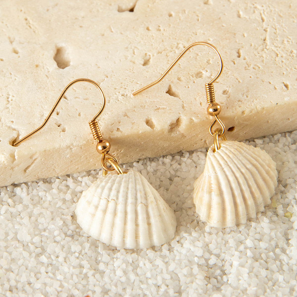 White scallop shell earrings with gold hooks placed on sand and stone background.
