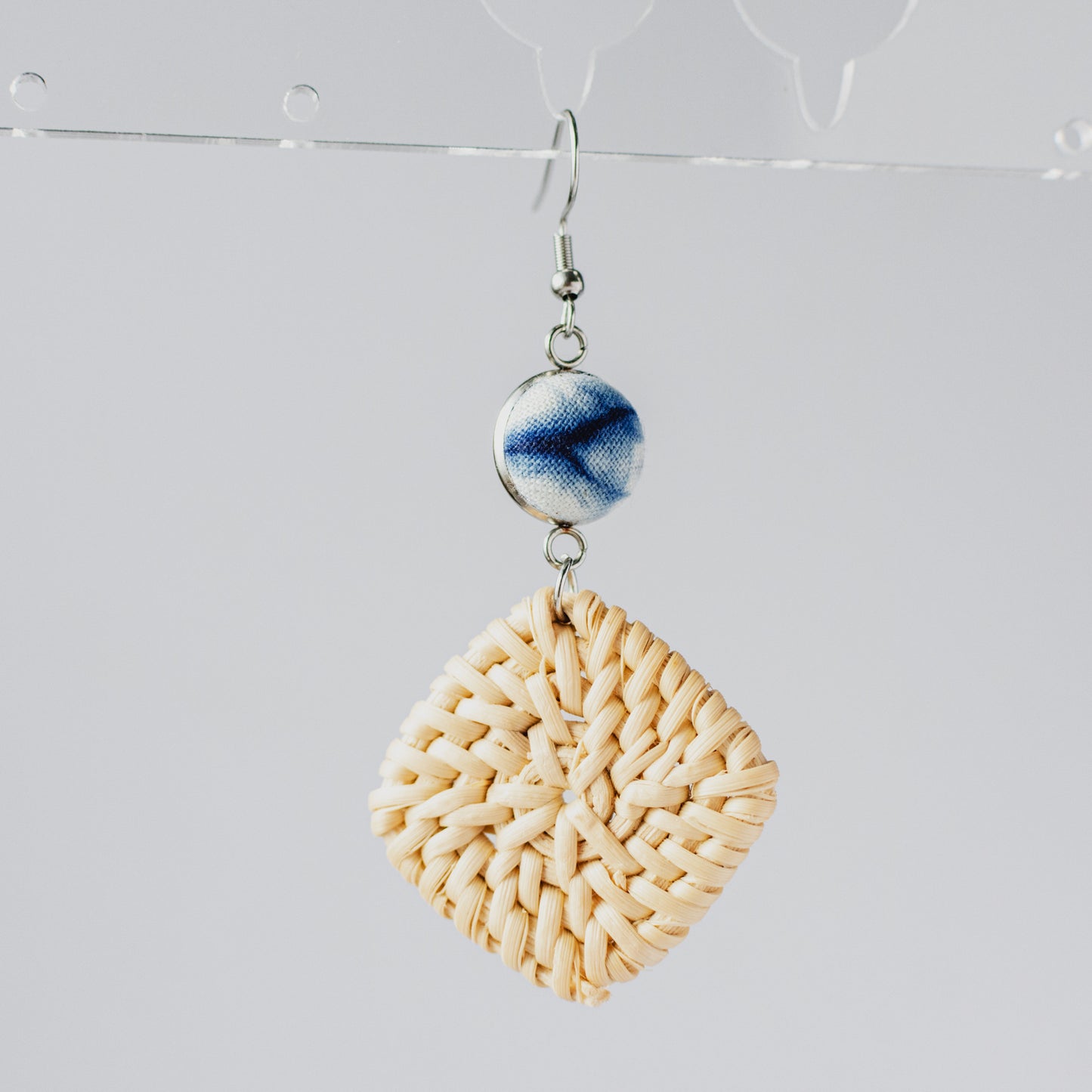 Single square woven straw earring with indigo-blue ceramic bead on white background.
