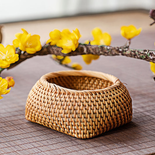 Small handmade rattan storage box with round-square shape placed on a bamboo mat, surrounded by yellow plum blossoms.