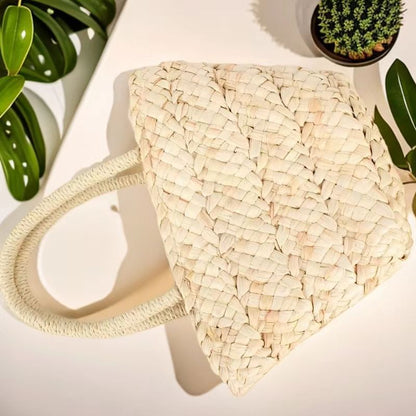 “Top-down view of handwoven corn husk handbag placed on white surface with green plants, by Solmerea.”