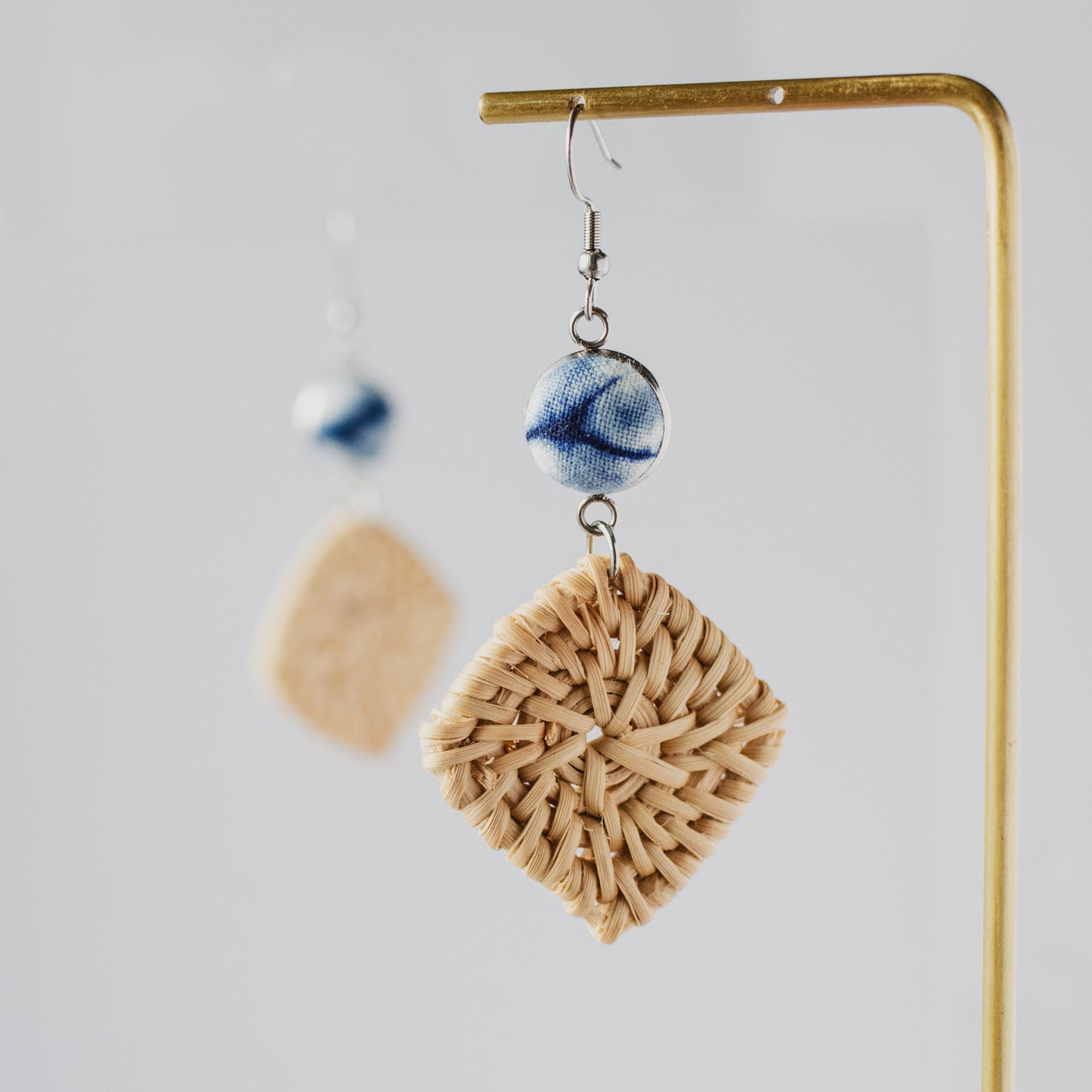 Close-up of square woven straw earring with blue ceramic bead on brass display stand.