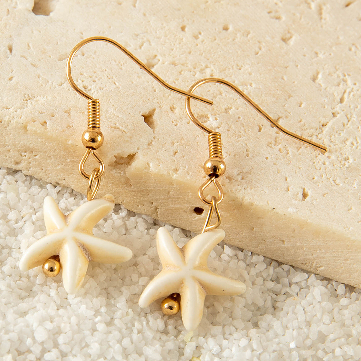 Close-up of gold hook starfish earrings resting on sand-colored stone surface.