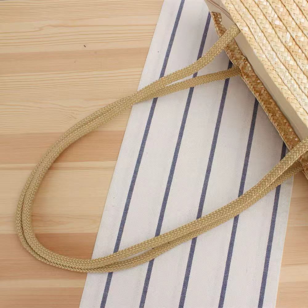 Handwoven straw beach tote bag standing upright against beige linen background, showing natural texture and simple silhouette.
