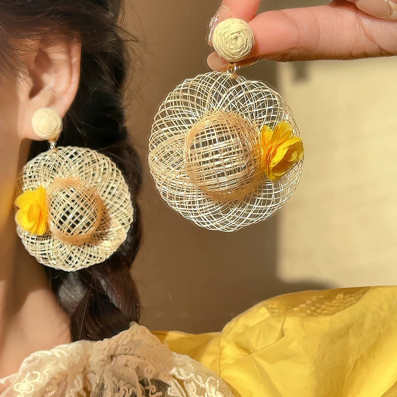 Woman wearing handwoven straw hat earrings with yellow flower accent under warm sunlight.