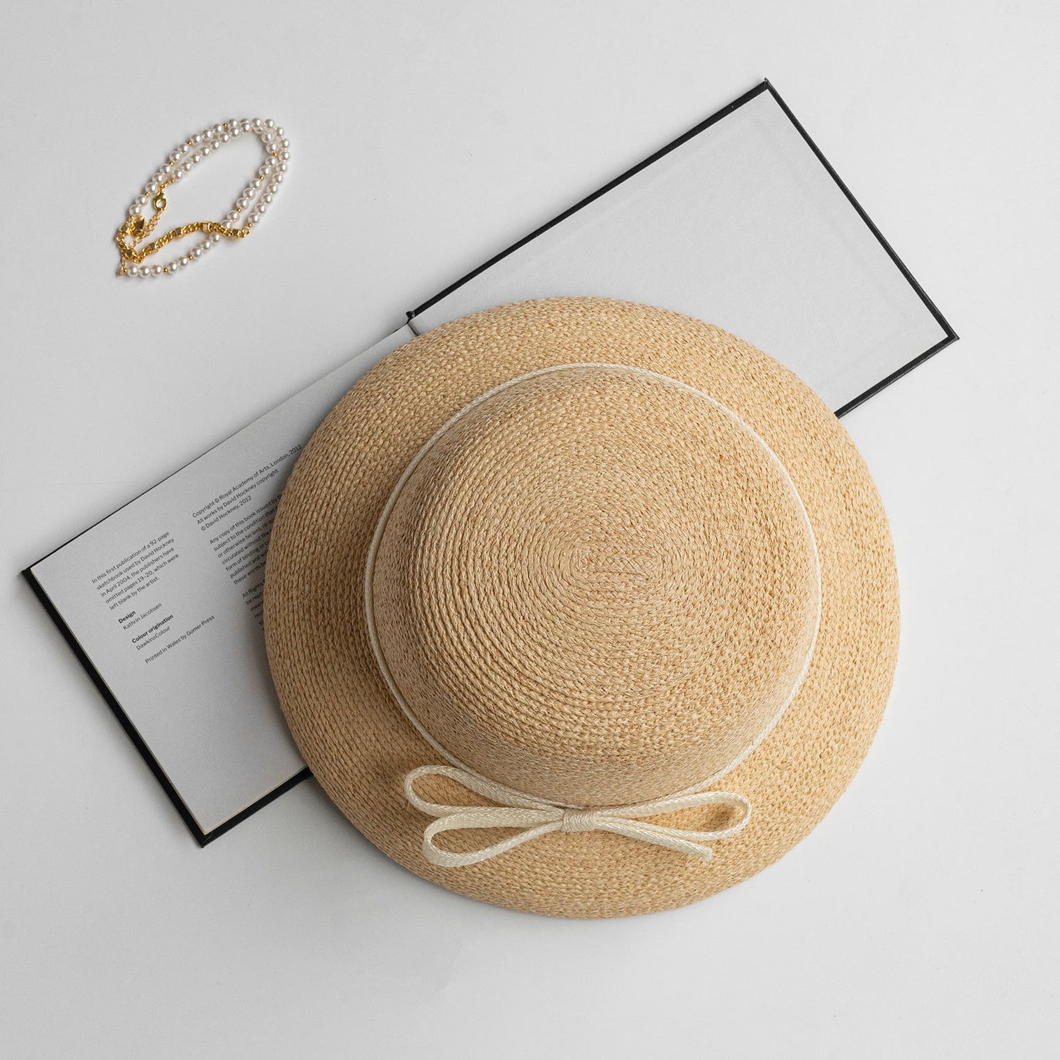 Top-down view of straw hat with cream bow, styled with pearl bracelet and open book for elegant flat-lay scene.