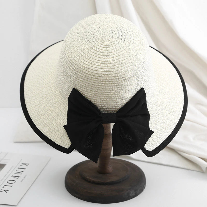 White straw hat with black edge trim and oversized bow, displayed elegantly on wooden stand.